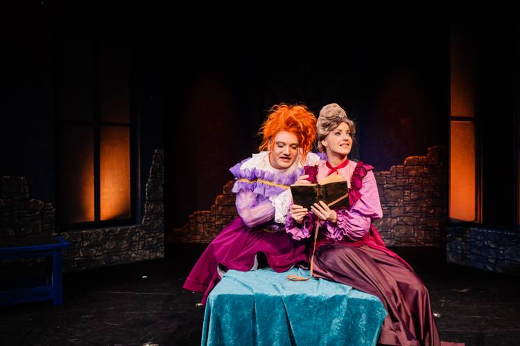Dracula – A Comedy of Terrors – Zwei Personen in viktorianischer Kleidung auf Bühne, halten geöffnetes Buch, sitzen auf blauem Tuch, dunkler Hintergrund mit steinernen Mauern.
