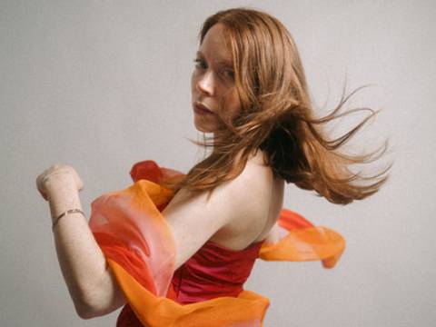 Laura Grandy – Laura Grandy mit langen rötlich-braunen Haaren in einem roten trägerlosen Kleid, umgeben von einem orangefarbenen, fließenden Stoff, der sich durch die Bewegung um sie legt. Sie blickt über die Schulter in die Kamera vor einem hellen, schlichten Hintergrund.