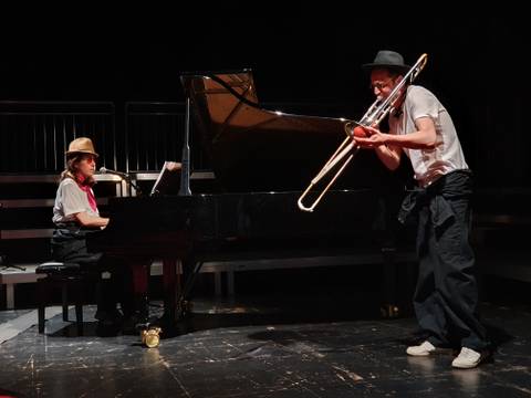Knirpskonzerte – Zwei Personen mit Hüten auf Bühne; eine spielt Klavier, die andere Posaune; auf dem Klavier liegt eine Partitur.