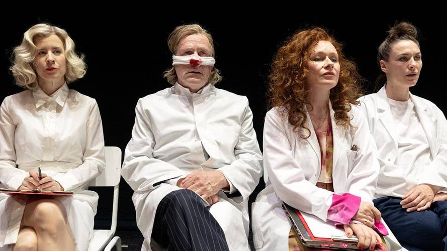 Vier Personen in weißen Kitteln sitzen auf Stühlen. Die mittlere Person hat verbundenen Kopf, die rechts daneben trägt violette Kleidung unter dem Kittel.