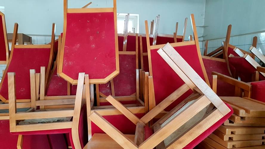 Stühle des Zentralen Runden Tisches (rechts) und Polstersessel der Volkskammer (links) in Einlagerungssituation vor 2019 – Rote Stühle mit Holzrahmen sind in einem Raum gestapelt. Die Stühle sind kopfüber aufeinander gestellt.