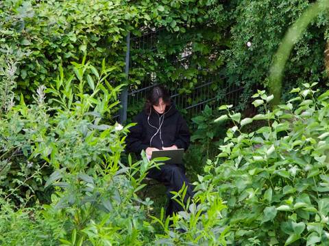  – Person mit Kopfhörern und Laptop sitzt im üppigen grünen Gebüsch, umgeben von dichtem Laub und Pflanzen.