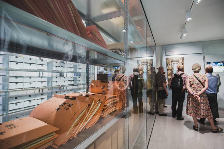 Personengruppe betrachtet Ausstellungsstücke. Links Regale mit Aktenordnern und beschrifteten Mappen. Dokumentarische Fotografie in einem Museum.