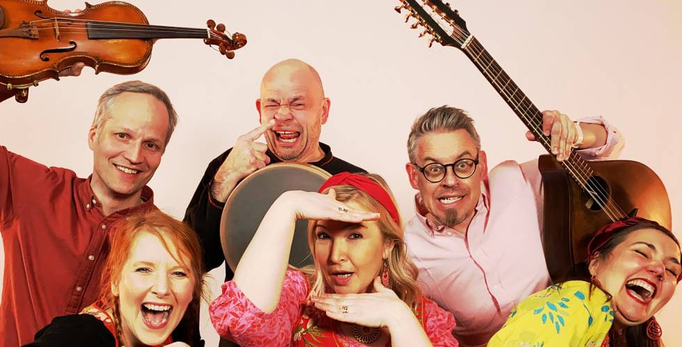Sechs Personen, lächelnd und posierend mit Instrumenten: Geige, Tamburin, Gitarre. Helle Kleidung, fröhliche Stimmung. Eine Person mit Brille.
