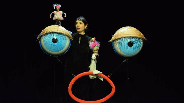 Die Wimpernzwerge – Person in schwarzem Outfit zwischen zwei großen blauen Augen. Rotes Accessoire im Hintergrund. Puppen auf Augen und in der Hand der Person.