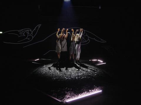 Bild 1: Aufführung des Stücks "Luftmasse" von Paper Tiger Theater Studio beim Theater der Welt Festival in Chemnitz – Vier Personen auf dunkler Bühne, Arme nach oben gestreckt. Leuchtende Linien erinnern an Hände im Hintergrund. пилынпьщму