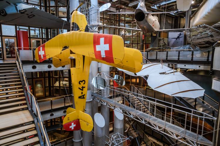 Die Bücker Bü 131 ist eines der ersten und weltweit bekanntesten Kunstflugzeuge. – Ein großes gelbes Flugzeug hängt senkrecht im offenen Treppenhaus des Neubaus. Im Hintergrund hängen weitere Flugzeuge und Flugapparate.