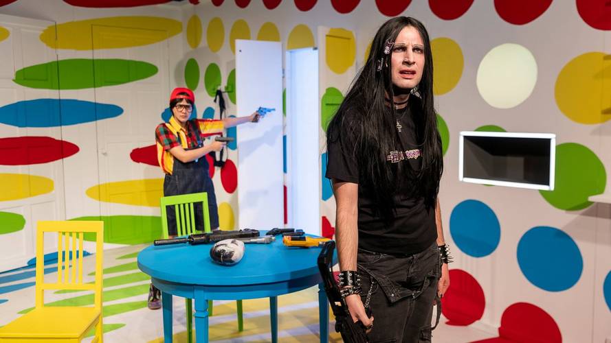 Zwei Personen in einem Raum mit bunten Punkten. Eine Person im Hintergrund mit Mütze und Spielzeugpistole. Tisch mit Waffen und Schädel.