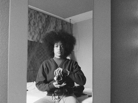 Nach, Selbstportrait – Schwarz-Weiß-Foto: Person mit Afro hält Kamera in Selbstporträt vor Spiegel. Person sitzt auf Bett, trägt Pullover und Jeans.
