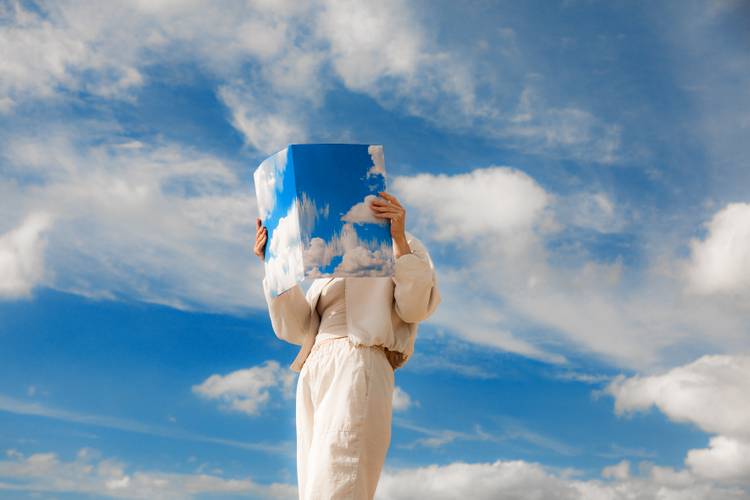 Person in hellem Outfit haelt ein großes Foto eines Himmels mit Wolken vor dem Gesicht, das sich nahtlos in den echten blauen Himmel mit Wolken einfügt.