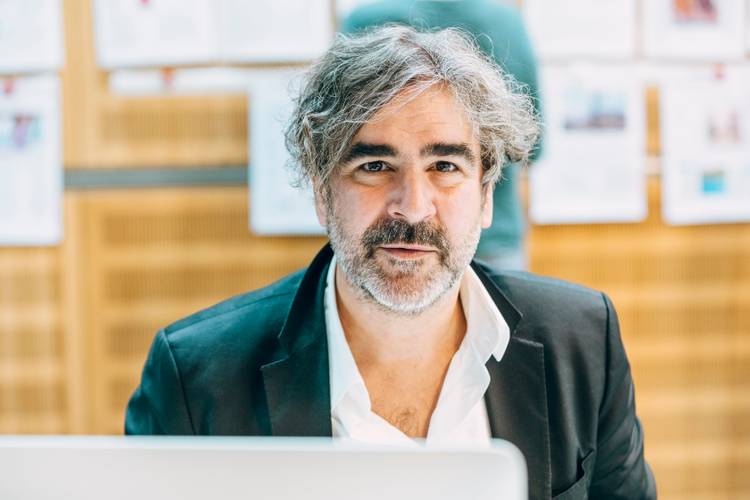 Deniz Yücel – Männlich gelesene Person mit grauen Locken und Bart, helle Haut, schwarzes Sakko, weißes Hemd, sitzt vor unscharfem Hintergrund mit Dokumenten.