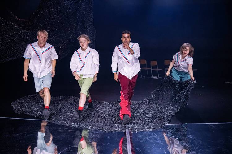 "Die Fische" am Theater Strahl – Vier Personen auf der Bühne ziehen ein großes schwarzes Netz, tragen weiße Hemden mit bunten Streifen. Im Hintergrund vier leere Stühle.