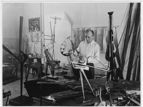 Ewald Gnilka, Ohne Titel (Hans Uhlmann in seinem Atelier), um 1954 – Schwarz-Weiss-Fotografie von Hans Uhlmann in seinem Atelier. Um den Künstler herum sind viele Metall-Skulpturen im Entstehungsprozess zu sehen.