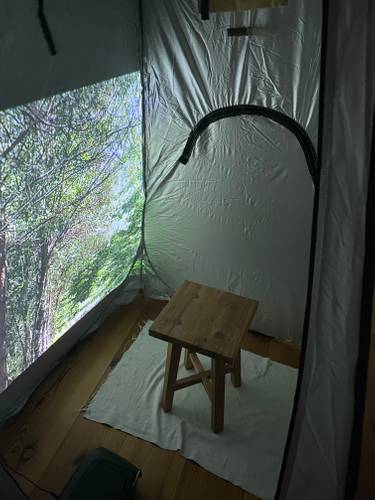 Einfach mal nichts tun. – Das Bild zeigt einen kleinen zeltartigen Raum mit silbernen Wänden. An eine Wand ist ein Foto von grünen Bäumen projeziert. In der Mitte des Raumes steht ein Hocker aus Holz auf einem weißen Teppich.