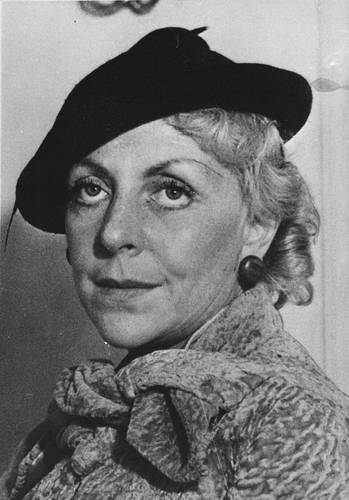 Vicki Baum – Person mit dunklem Barett und hellem, texturiertem Mantel. Kurze, wellige Haare. Die Person blickt ernst. Hintergrund unscharf.