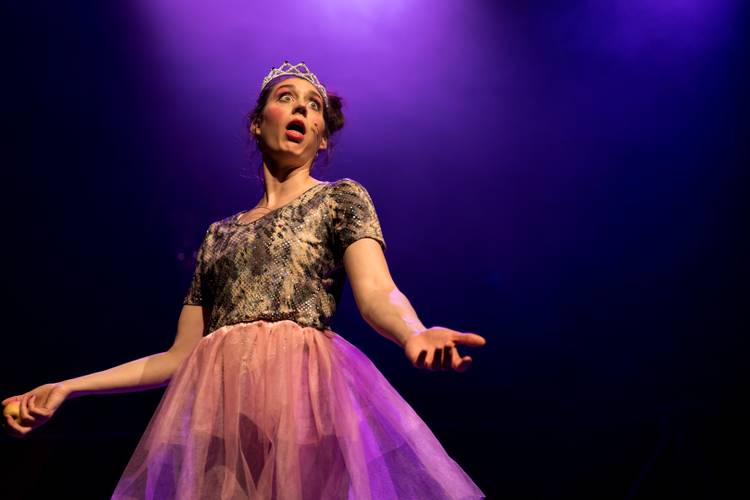 Barbaren Barbies: A wild women circus – Person mit Krone und buntem Kleid auf Bühne, dramatische Pose, lila beleuchtet, weit geöffneter Mund, dunkler Hintergrund.