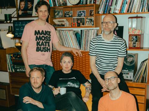 Elin Forkelid – Gruppe von fünf Personen in einem Wohnzimmer mit Bücherregalen, Schallplatten und einem auffälligen T-Shirt in Rosa.