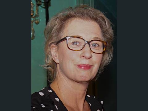  – Eine Person mit Brille und kurzen Haaren trägt ein schwarz-weiß gepunktetes Kleid und lächelt leicht. Hintergrund grünlich.