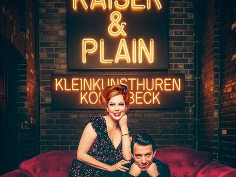 KAISER & PLAIN – Zwei Personen auf rotem Sofa, eine im glitzernden Kleid, eine in dunkler Kleidung. Leuchtschrift: KAISER & PLAIN, KLEINKUNSTHUREN KOTTBÄCK.