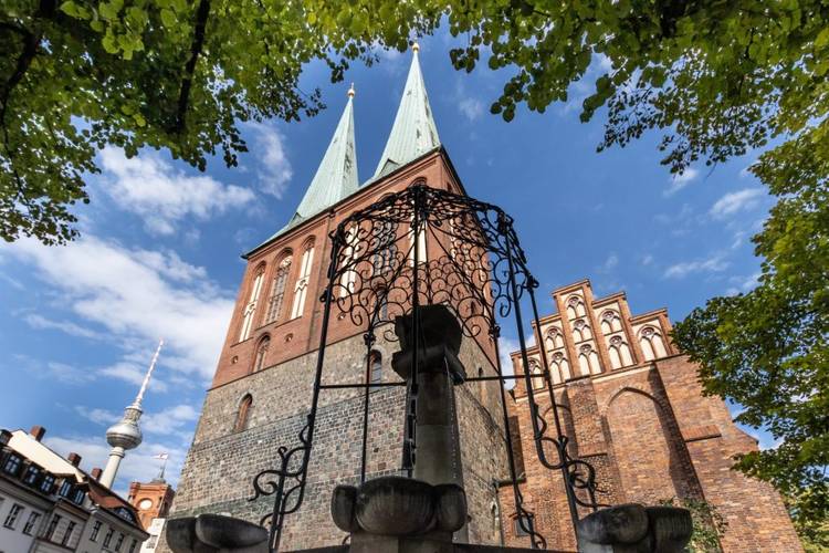 Das Museum Nikolaikirche ist nicht weit von weiteren geschichtsträchtigen Orten Berlins entfernt: dem Rote Rathaus und dem Fernsehturm. – lick durch eine schmiedeeiserne Brunnenzier auf die Türme und Giebel der Nikolaikirche, im Hinterfgrund hinter den Dächern historisierender Gebäude der Fernsehturm und der Turm des Roten Rathauses