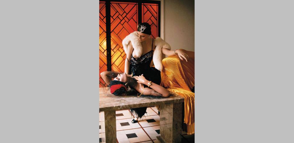 Helmut Newton, Store Dummies, Paris 1976 – Zwei Personen in glamouröser Kleidung auf einem Marmortisch. Eine trägt ein gelbes Kleid, die andere eine schwarze Robe mit Pelz. Hintergrund geometrische