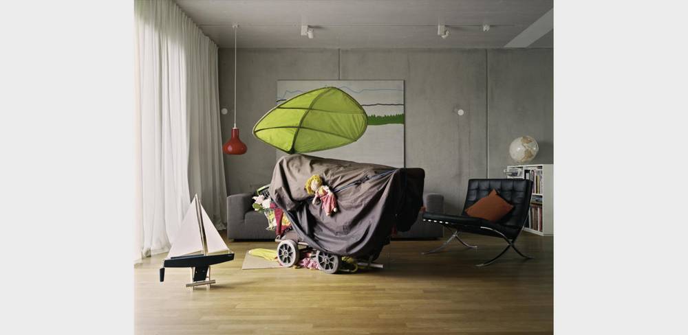 Wohnzimmer mit Spielzeugboot, abgedecktem Kinderwagen, grünem Blätterdach. Moderne Möbel, rote Hängelampe, ungewisse witterung. Wandbild im Hintergrund.