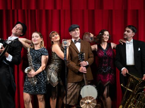  – Sechs Personen in Vintage-Kleidung vor rotem Vorhang: mit Gitarre, Saxophon, Mikrofon, Ukulele, Banjo, Tuba. Links stehender Hutträger in Anzug.
