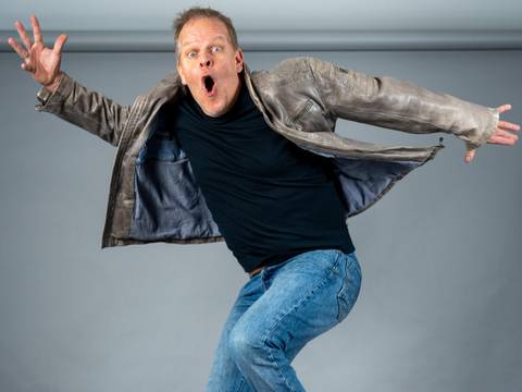  – Eine Person in blauer Jeans und schwarzem T-Shirt mit geöffneter Lederjacke springt mit ausgebreiteten Armen und offenem Mund.