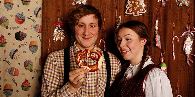 Person mit kariertem Hemd haelt Brezel; daneben weiblich gelesene Person mit Zopf, laechelnd. Hintergrund mit Lebkuchen und Text: Sweet, Cupcake, Sugar.