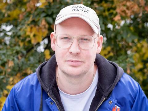 Tim Holland – Person mit Brille und Kappe mit Aufschrift "THE NAME IS POETRY", in blauer Jacke mit Basketball-Logo, vor unscharfem Hintergrund mit Bäumen.
