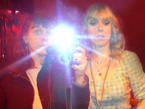Sandra Grether und Kersty Grether – Zwei Frauen stehen vor einem Spiegel, eine hält eine Kamera in der Hand und nimmt dieses Portrait der beiden mit einem Blitzlicht auf