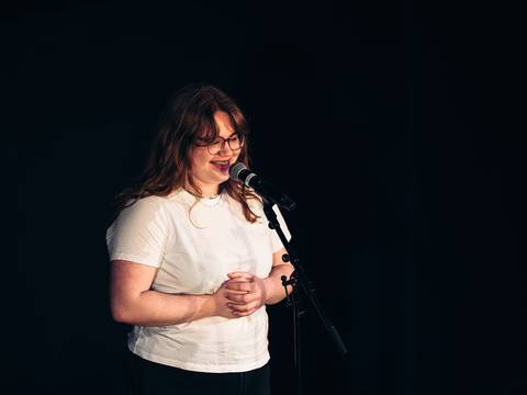  – Eine lächelnde Person mit Brille und weißem T-Shirt steht vor einem Mikrofon. Dunkler Hintergrund.