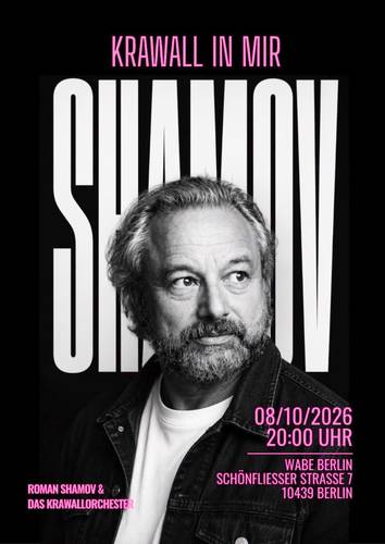 Schwarzweiß-Porträt von Roman Shamov mit mittellangem, grauem Haar und Vollbart. Roman Shamov blickt leicht nach rechts. Im Hintergrund befinden sich vertikale schwarze Streifen auf hellem Grund. Die Aufnahme zeigt sein Gesicht und den oberen Schulterbereich und wirkt kontrastreich sowie professionell fotografiert.