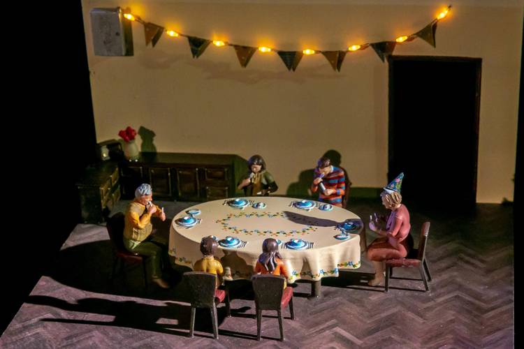 Transport: Departure – Acht Miniaturfiguren sitzen um einen runden Tisch mit gelber Tischdecke, gedeckt mit blauem Geschirr. Lichterkette und Wimpel im Hintergrund.
