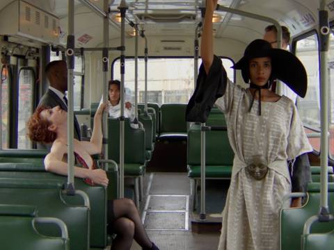 Samuel Chalela Puccini – Person mit schwarzem Hut und hellem Kleid steht in einem Bus und hält sich an der Stange fest. Andere Personen sitzen auf grünen Sitzen.