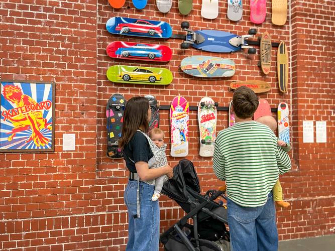 ElternZEITgeschichte – Zwei Frauen stehen vor einer Backsteinwand und betrachten Skateboards an der Wand. Beide Frauen tragen Babys auf dem Arm, im Hintergrund steht ein Kinderwagen.
