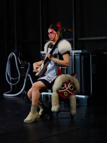 Foto: Diana Pfammatter – Person mit roten Gesichtsbemalungen spielt ein Saiteninstrument, trägt Fellbesatz und sitzt neben einer Maske mit roten Ohren und gelben Augen.