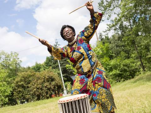 Traditional dance from Guinea – Person in buntem Kleid trommelt im Freien. Bäume und Wiese im Hintergrund. Energische Pose mit hoch erhobenen Trommelstöcken.