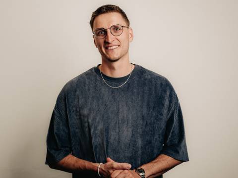 Johannes Steuding – Eine Person mit Brille und kurzem Haar trägt ein dunkelblaues Oberteil und eine Kette, lächelt vor hellem Hintergrund.