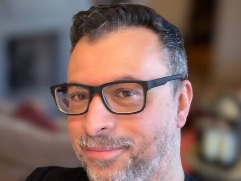Fabrice Sapolsky – Person mit Brille und Bart, trägt ein schwarzes T-Shirt, sitzt lächelnd. Hintergrund unscharf.