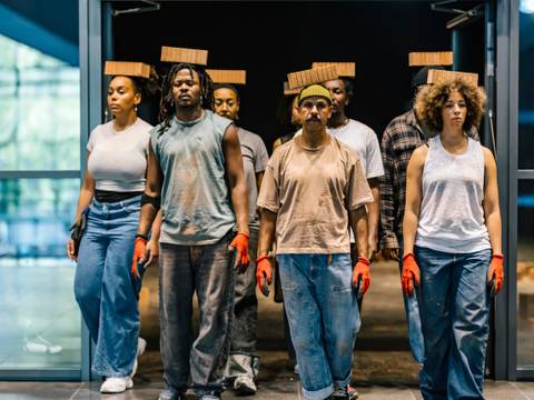 Aufführung des Stücks "ON MY WAY HOME" von Reinaldo Reibeiro und Lucia Oiro im Rautenstrauch-Joest-Museum in Köln – Sechs Personen tragen Backsteine auf dem Kopf. Alle tragen rote Handschuhe, Jeans und helle Oberteile. Sie stehen in einem Eingangsbereich.
