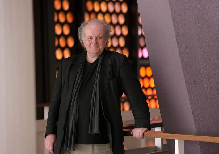 Wolfgang Rihm – Ältere, männlich gelesene Person mit schwarzem Schal und Jacke steht lächelnd an einem Holzgeländer. Hintergrund aus orange leuchtenden Kreisen.
