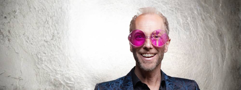 Michael Krebs: OPTIMISMUS – jetzt stark reduziert – Person mit rosa Brille und buntem Jackett vor hellem Hintergrund, lächelt in die Kamera.