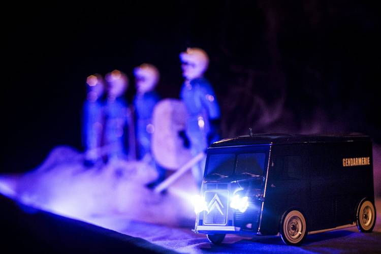 Blue – Spielzeuglieferwagen mit Aufschrift „Gendarmerie“ und leuchtenden Scheinwerfern. Im Hintergrund unscharfe Figuren in Uniformen mit Schwertern und Schilden.