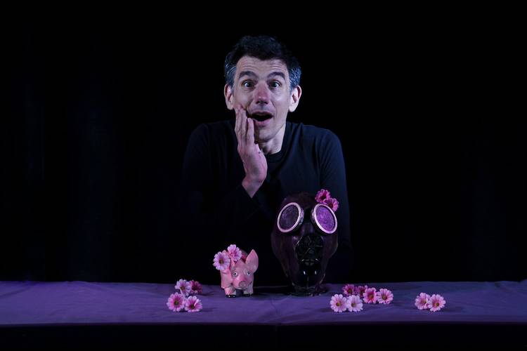 Blue – Mensch mit erstaunter Mimik, Maske mit rosa Gänseblümchen dekoriert, rosa Schweinchenfigur auf lila Tisch, darunter verstreute rosa Gänseblümchen.
