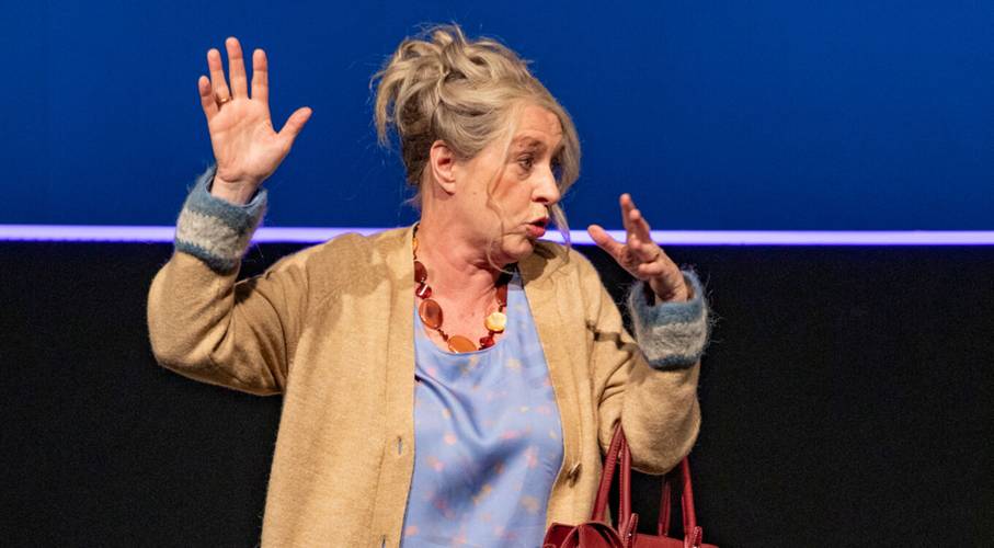 Was ist los mit der Welt?! – Ältere, weiblich gelesene Person mit beigem Cardigan und blauer Bluse gestikuliert mit erhobenen Händen vor blauem Hintergrund.