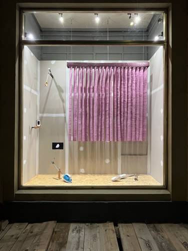 Künstlerinnengespräch »A house and two windows« – Schaufenster mit rosa Vorhang, Holzlöffel an der Wand, Bügeleisen, Fahrradschlüssel und ein Bild eines Eisbergs auf hellem Boden und Wand.