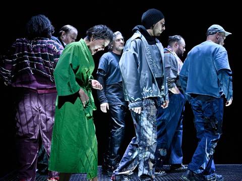  – Personengruppe auf einer Bühne, verschiedene bunte Outfits, darunter grüne und blaue Kleidung, auf schwarzem Hintergrund. Keine erkennbare Interaktion.