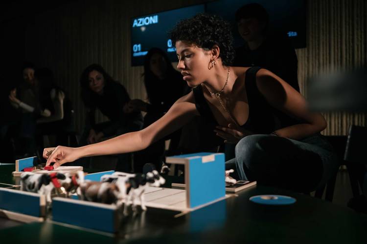 The Game – Person beugt sich über Tisch mit Miniaturmodellen von Kühen. Im Hintergrund sitzen Personen. Anzeige mit Text: AZIONI 01.