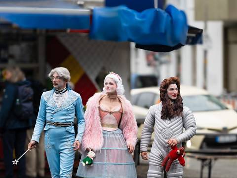 Muttis Kinder: Intergalactic Interruption – Drei Personen in auffälligen Kostümen gehen nebeneinander auf einer nassen Straße. Sie tragen Spielzeugtiere und schauen nach vorne.